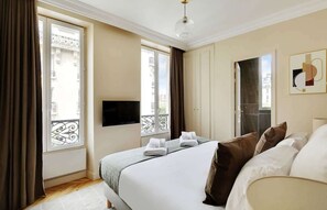 A hotel room with a large bed, a flat-screen TV mounted on the wall, and a balcony with a view of the city. - An Exceptional 3-BR/2Ba - Invalides (Paris)