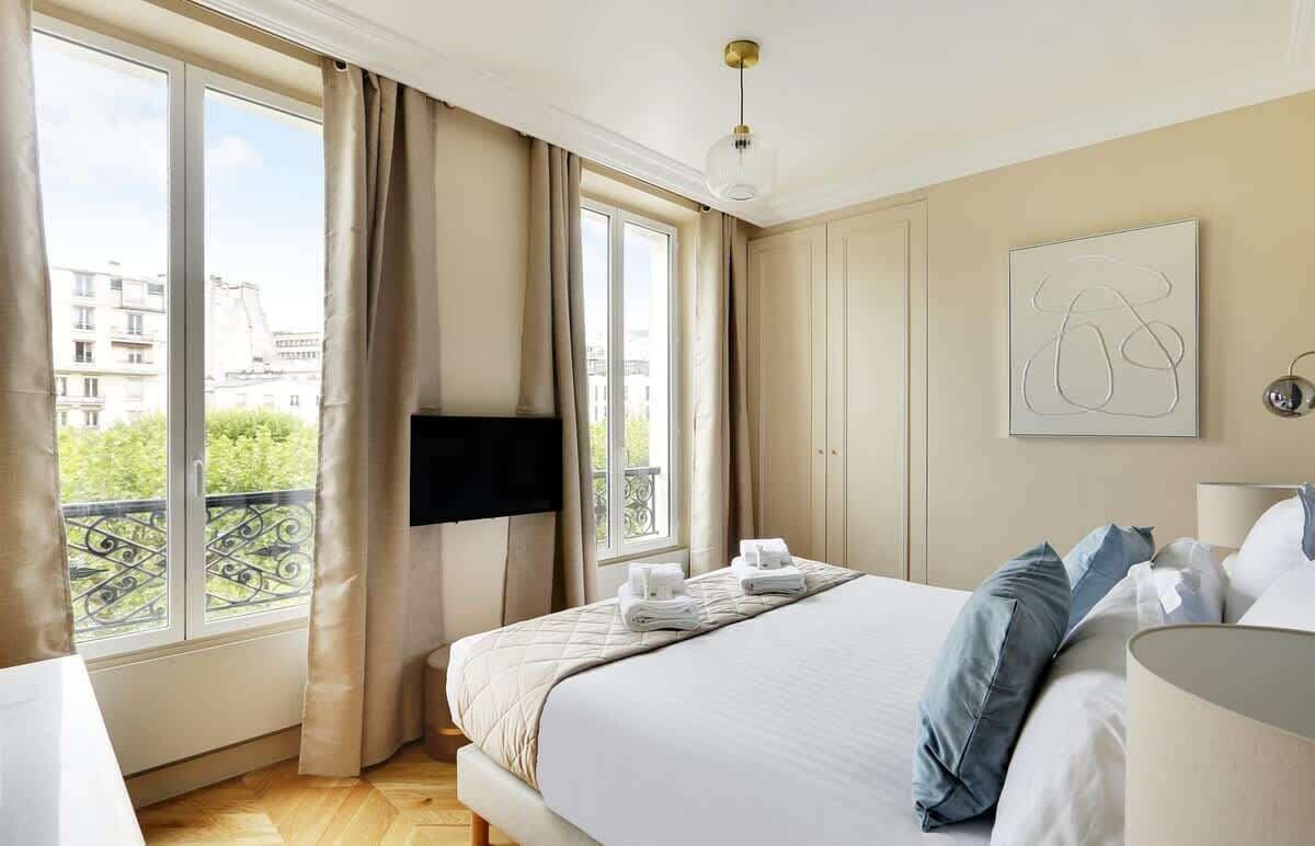 Habitación de hotel con una cama grande, un televisor de pantalla plana en la pared, una ventana con cortinas y un balcón con vistas a edificios y zonas verdes.