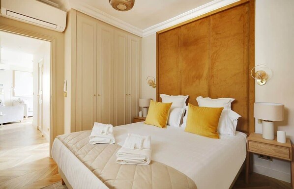 A hotel room with a large bed, wooden headboard, two bedside tables with lamps, and a door leading to another room. - An Exceptional 3-BR/2Ba - Invalides (Paris)