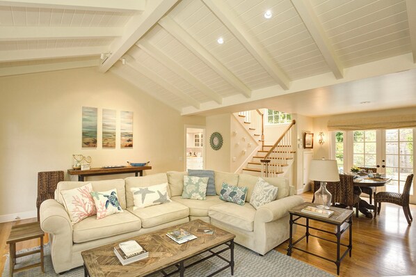 A spacious living room with a large sectional sofa, a coffee table, and a dining area with a table and chairs. A staircase with a railing is visible in the background.