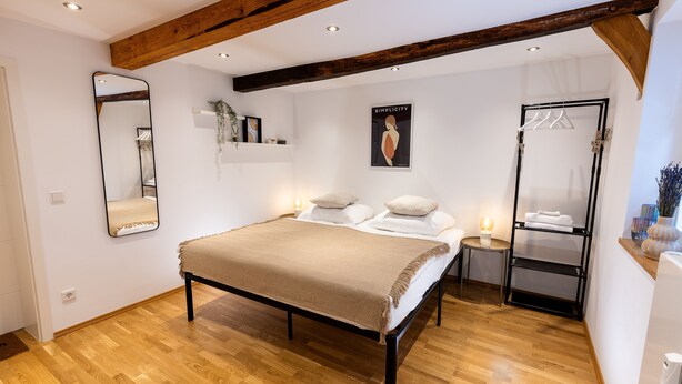 A bedroom with a wooden floor, a bed with a beige blanket, a bedside table with a lamp, a mirror, a poster on the wall, and a wardrobe with hanging clothes. - 1 Bedroom Apartment in Hattingen (Hattingen)