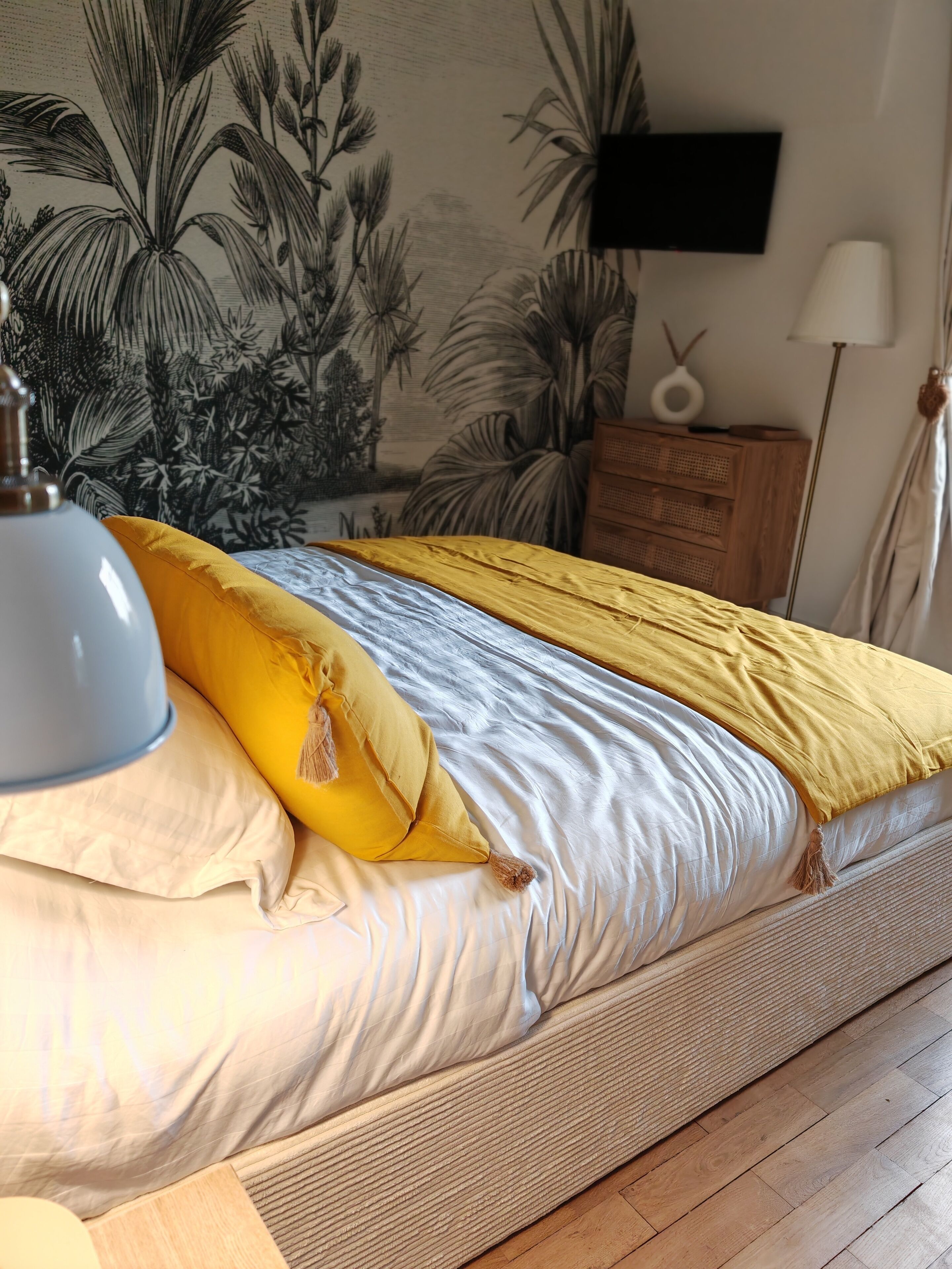 Un lit avec une literie blanche et jaune, une lampe, une commode en bois et un mur orné d’une fresque aux motifs tropicaux.
