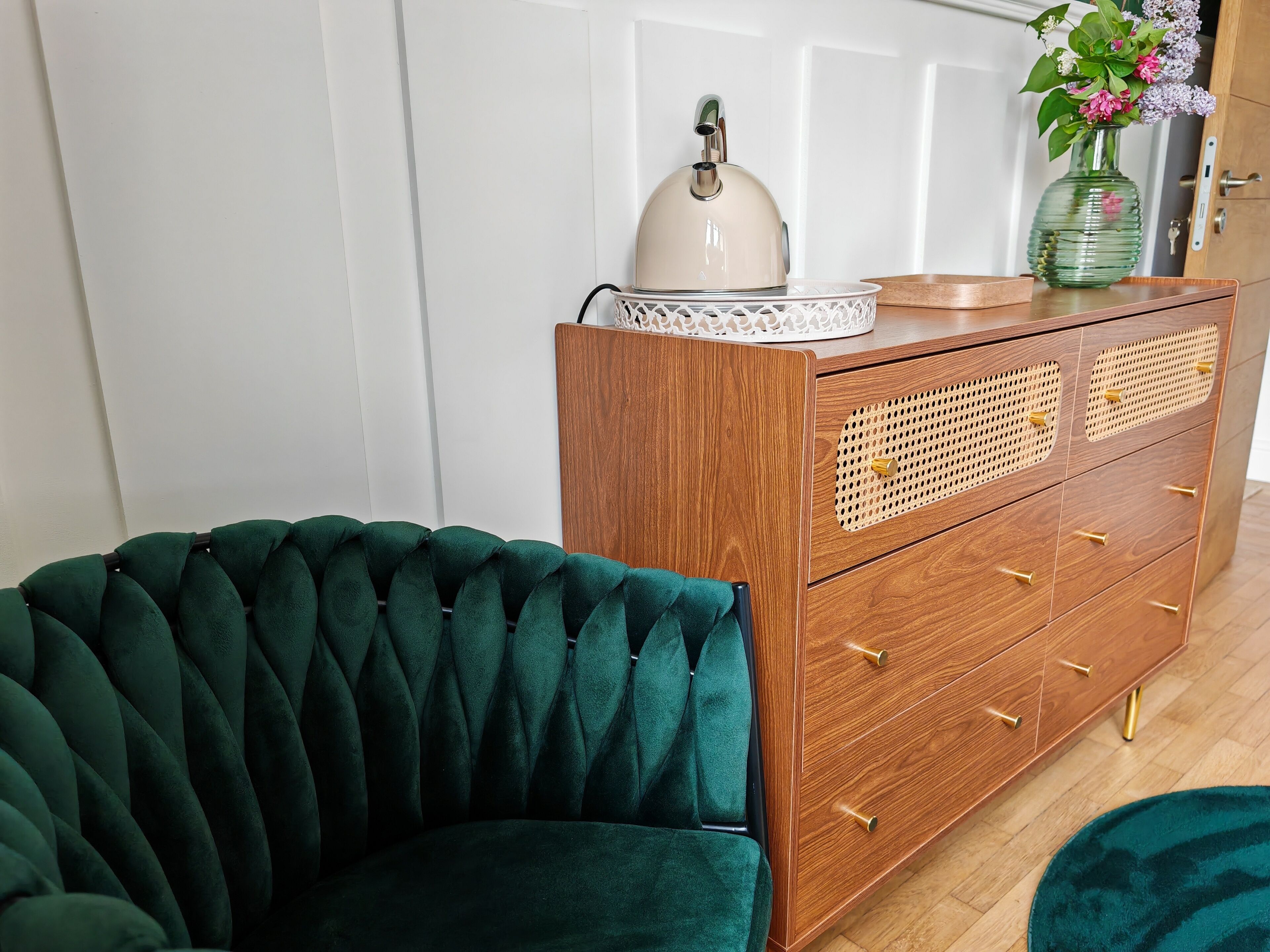 A room with a green velvet armchair, a wooden dresser with drawers, a white door, and a vase with flowers on top of the dresser.