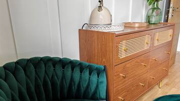 A room with a green velvet armchair, a wooden dresser with drawers, a white door, and a vase with flowers on top of the dresser.