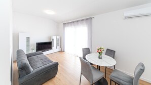 Ein modernes Wohnzimmer mit grauem Sofa, einem Flachbildfernseher, einem Glastisch und einem Essbereich mit Marmortisch und grauen Stühlen.