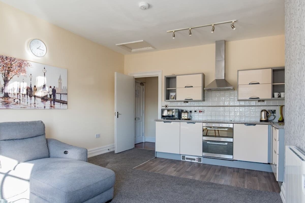 A modern apartment with a grey sofa, a kitchen with white cabinets, a built-in oven, and a clock on the wall.