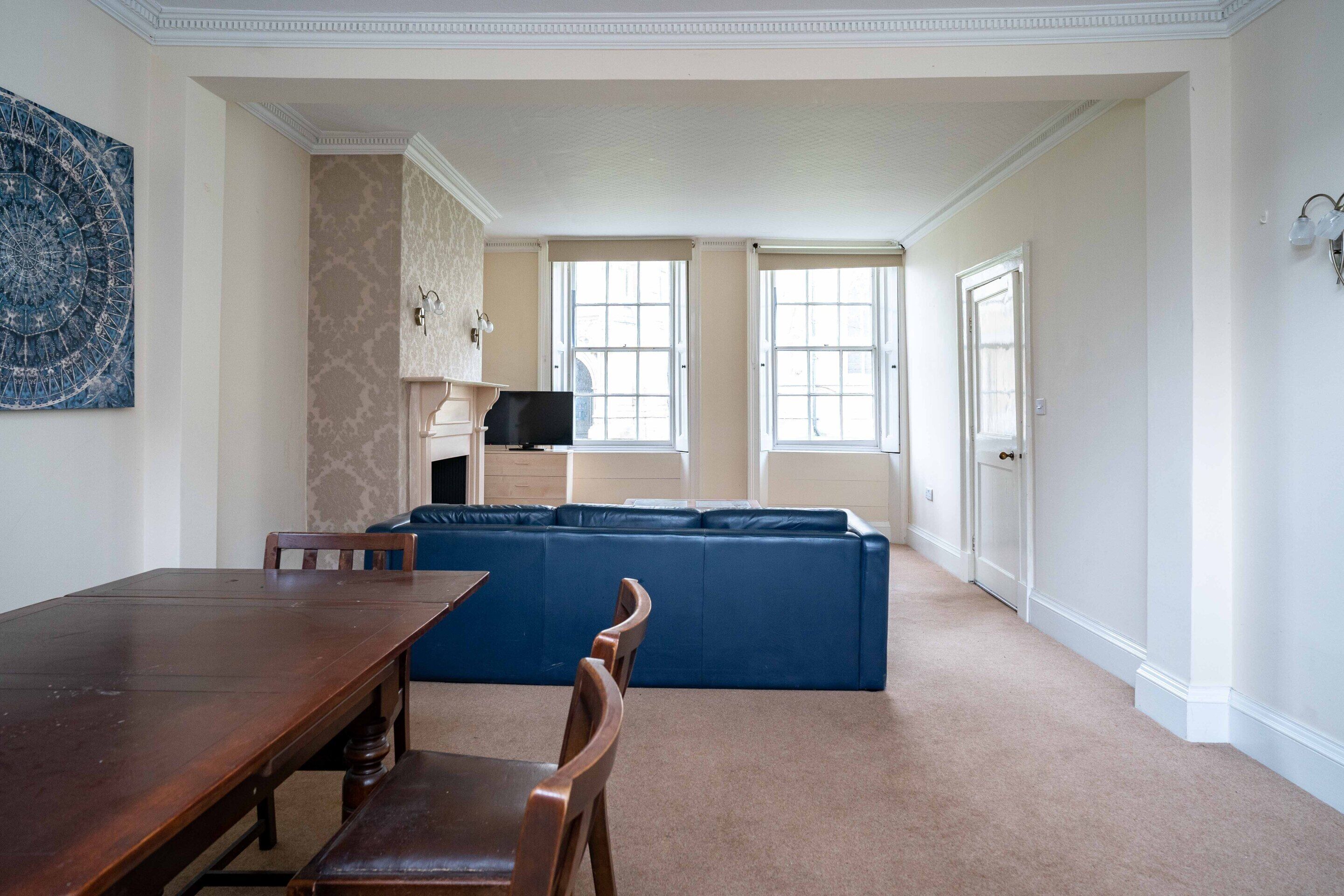 A spacious living room with a large blue sectional sofa, a wooden dining table, and a kitchen area in the background.
