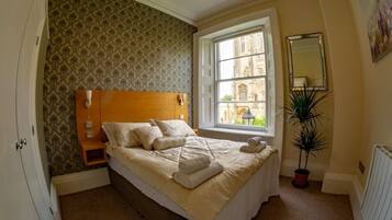 A neatly made bed with a headboard, a bedside table, a window with a view of a building, and a potted plant.