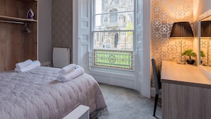 A bedroom with a large window, a radiator, a bed with rolled towels, a wooden shelf, and a desk with a lamp and a small plant. - Church Lane Apartments (Boston)
