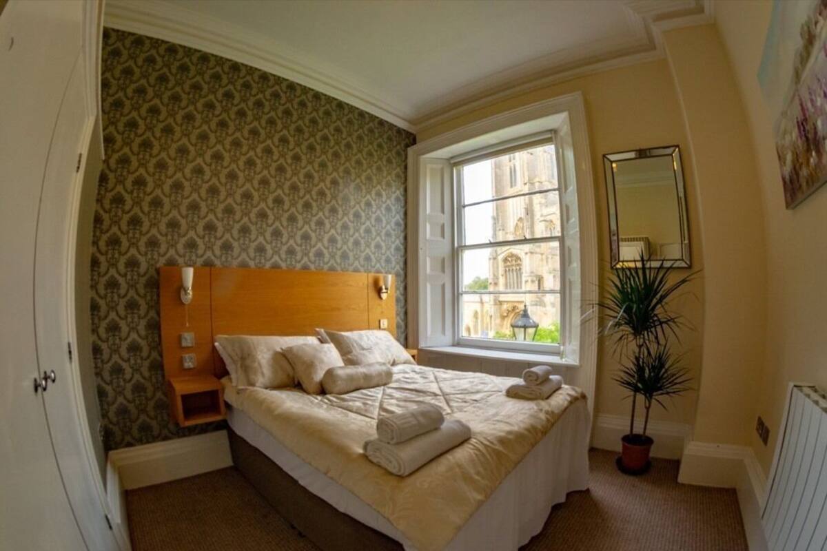 A neatly made bed with pillows and towels, a bedside table, a window with a view of a building, and a potted plant.