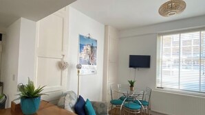 Un salon moderne avec une table à manger en bois, un canapé agrémenté de coussins turquoise, un téléviseur fixé au mur et une grande fenêtre avec des stores.