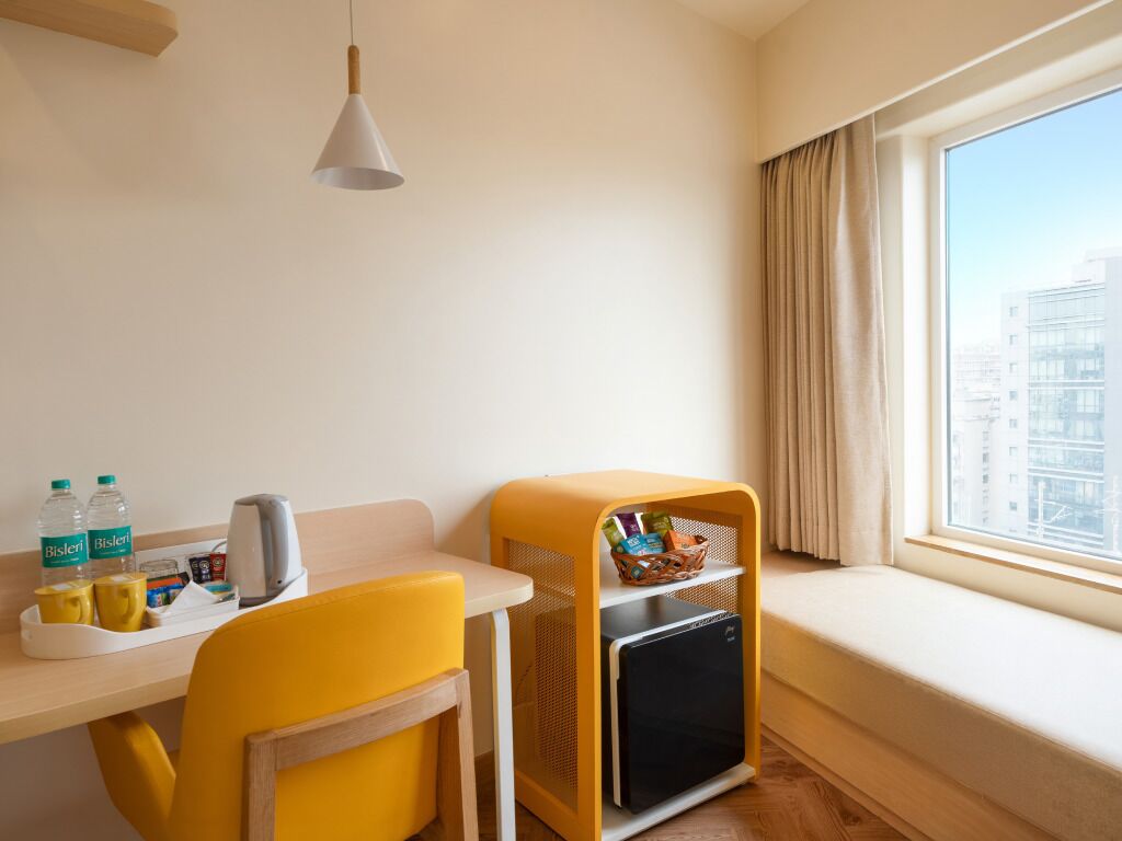 A hotel room with a small table, a yellow chair, a mini-fridge, a window with curtains, and a view of buildings outside.