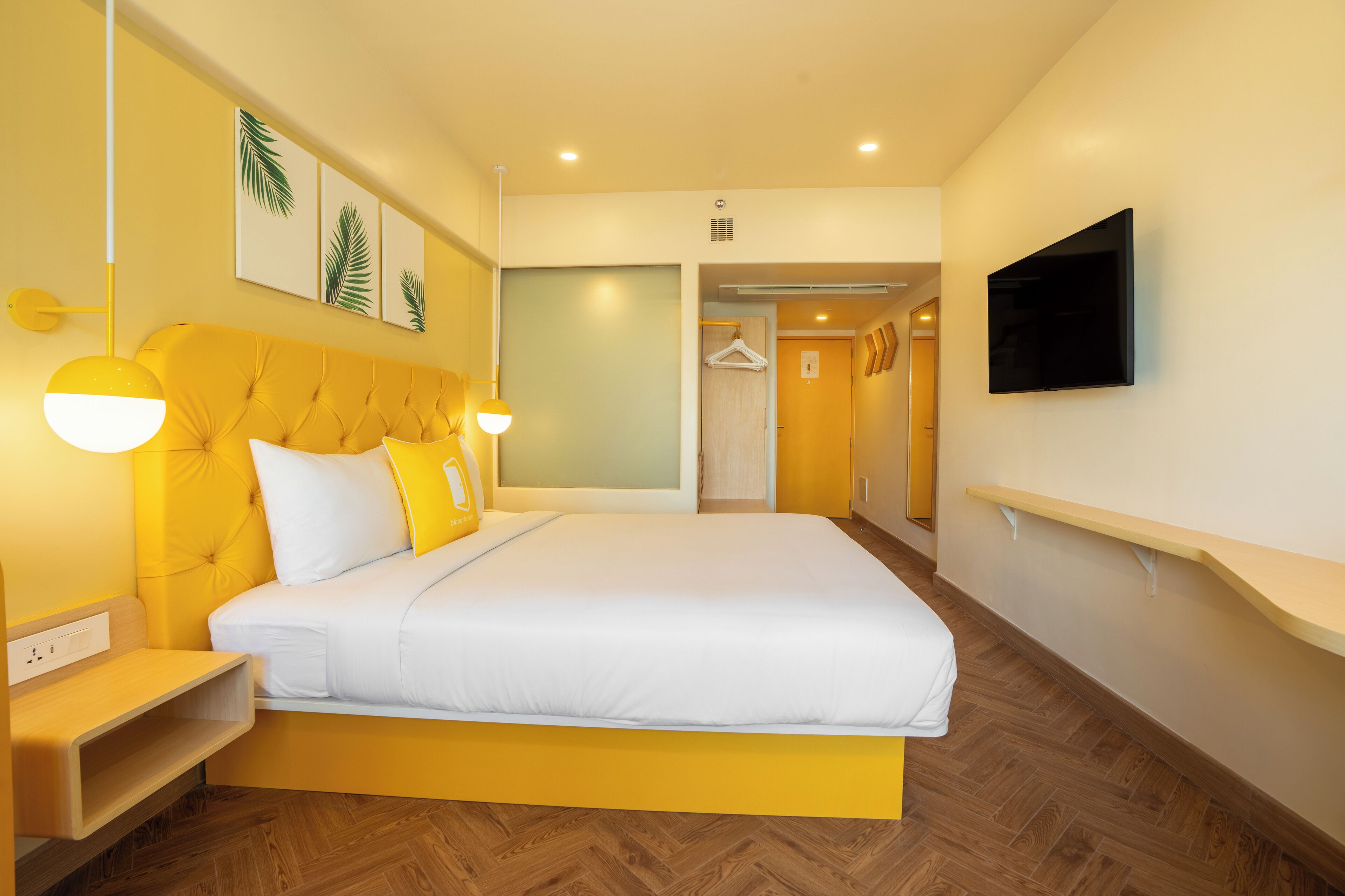 Una habitación de hotel moderna con una cama amarilla, ropa de cama blanca, un televisor montado y suelo de madera.