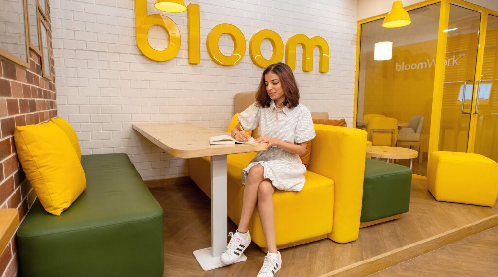 A person sitting at a table in a room with yellow and green furniture, a brick wall, and a 'bloom' sign on the wall.