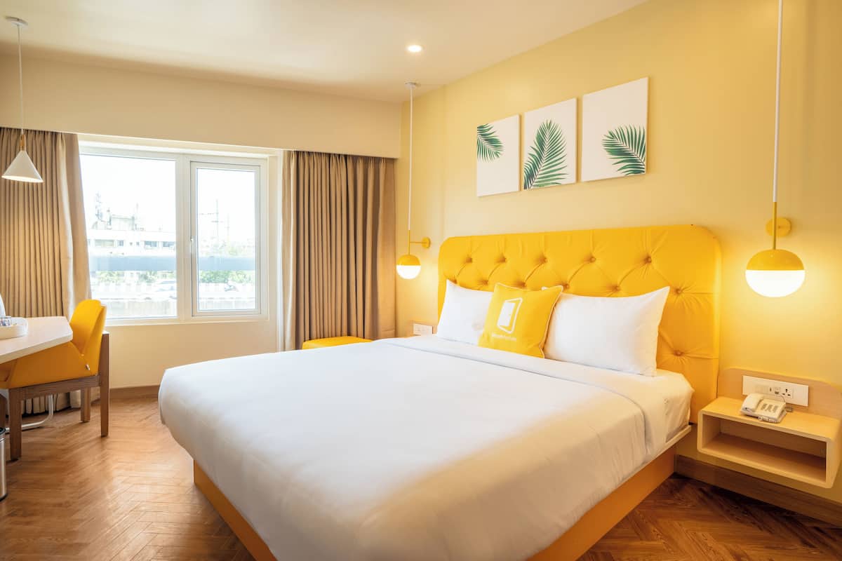 A hotel room with a large bed, a desk with a chair, a window with curtains, and a nightstand with a phone.