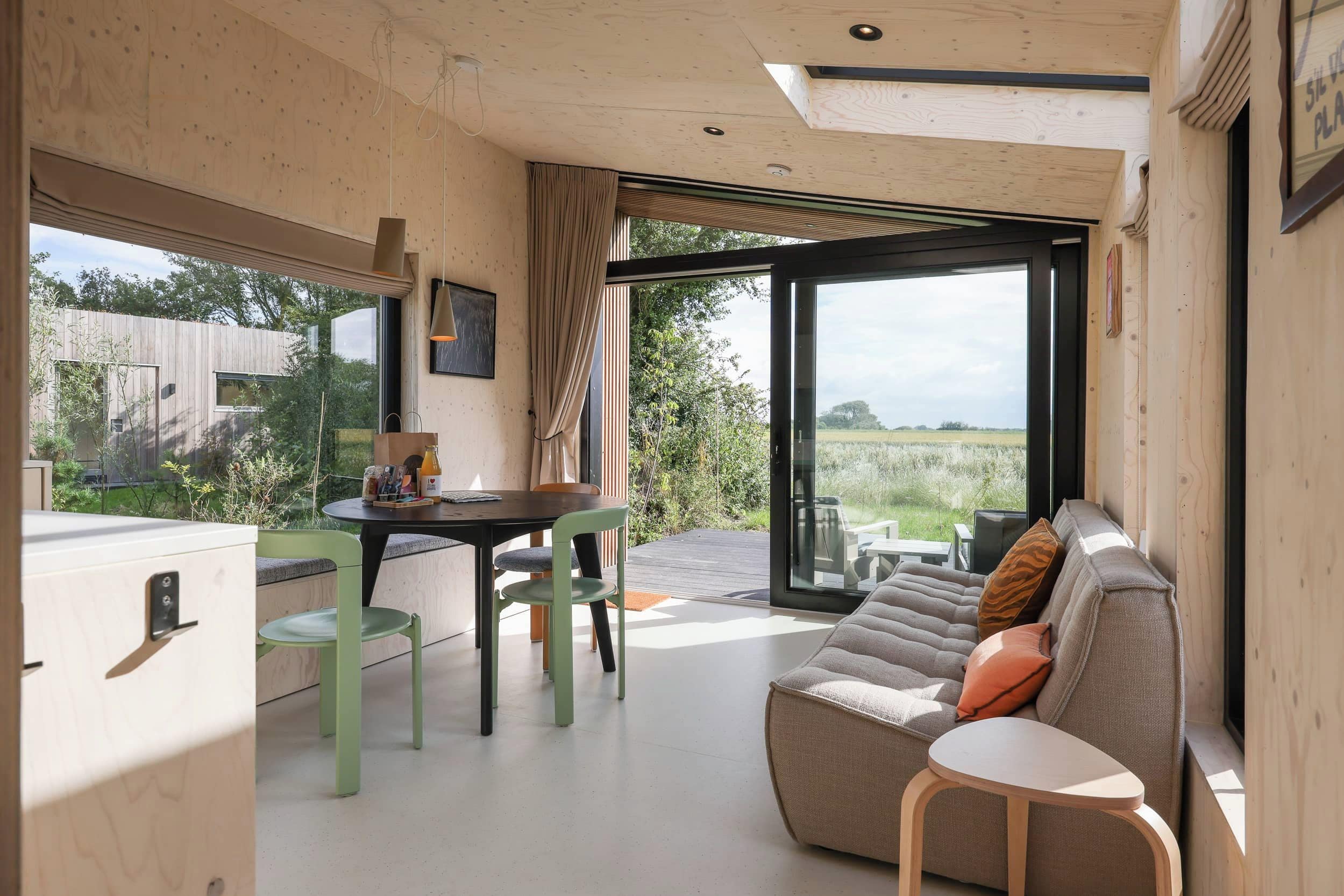 Un interior moderno con techo de madera, una zona de comedor con mesa y sillas, un sofá con cojines y un amplio ventanal corredero que da a un espacio exterior.