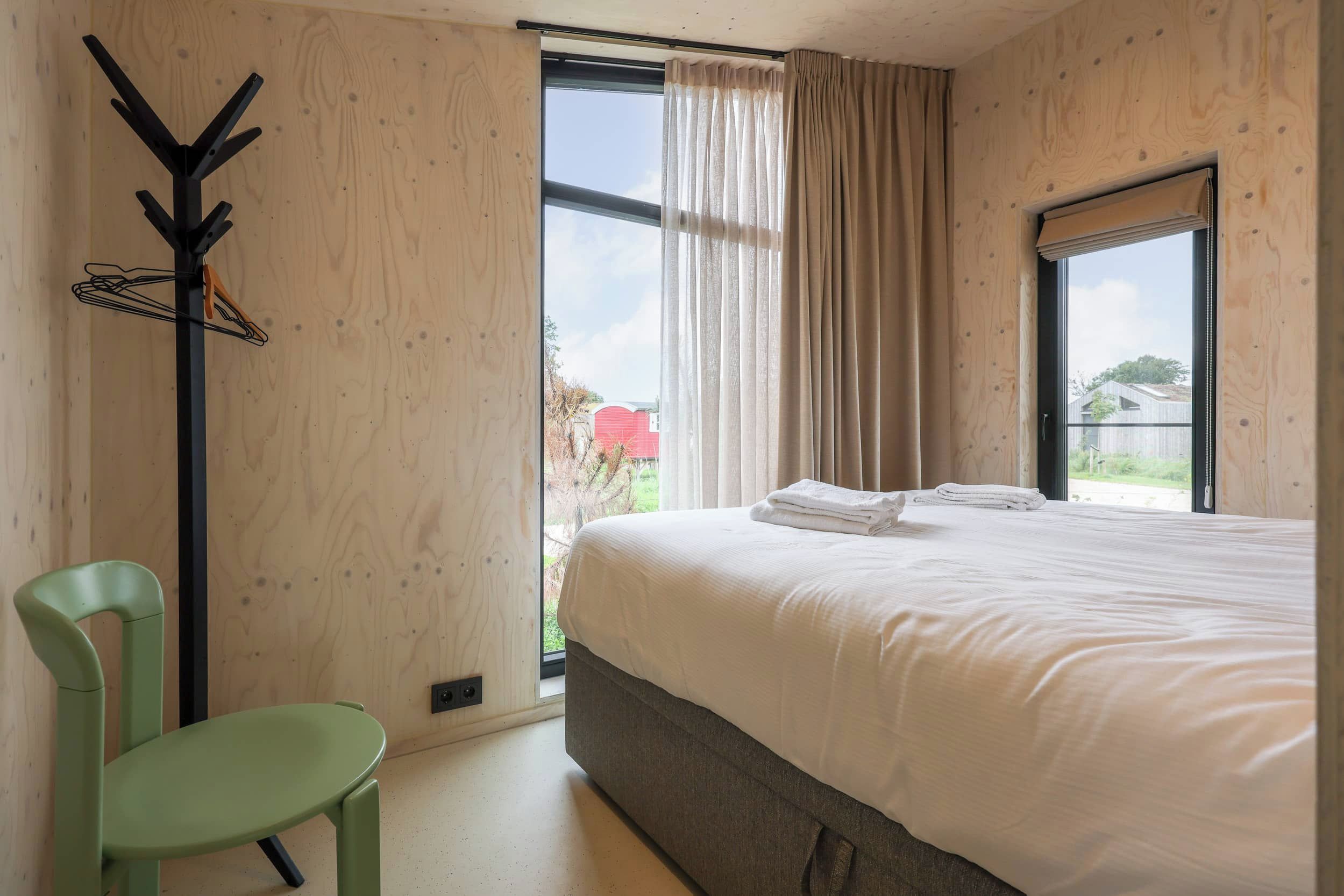 Ein Schlafzimmer mit Bett, Stuhl, Kleiderhaken und einem Fenster mit Vorhängen.