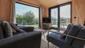 A modern living room with a sofa, armchair, and TV, large windows with a view of greenery, and a wooden ceiling.