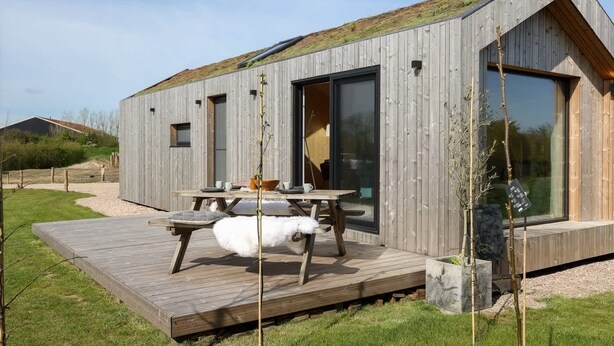 A modern wooden cabin with a deck, large windows, and a small outdoor seating area.