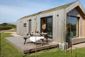 Una cabaña moderna de madera con terraza, ventanas amplias y un pequeño rincón de estar al aire libre.