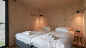 A double bed with white linens and towels, a small bedside table, and a view through a large window.