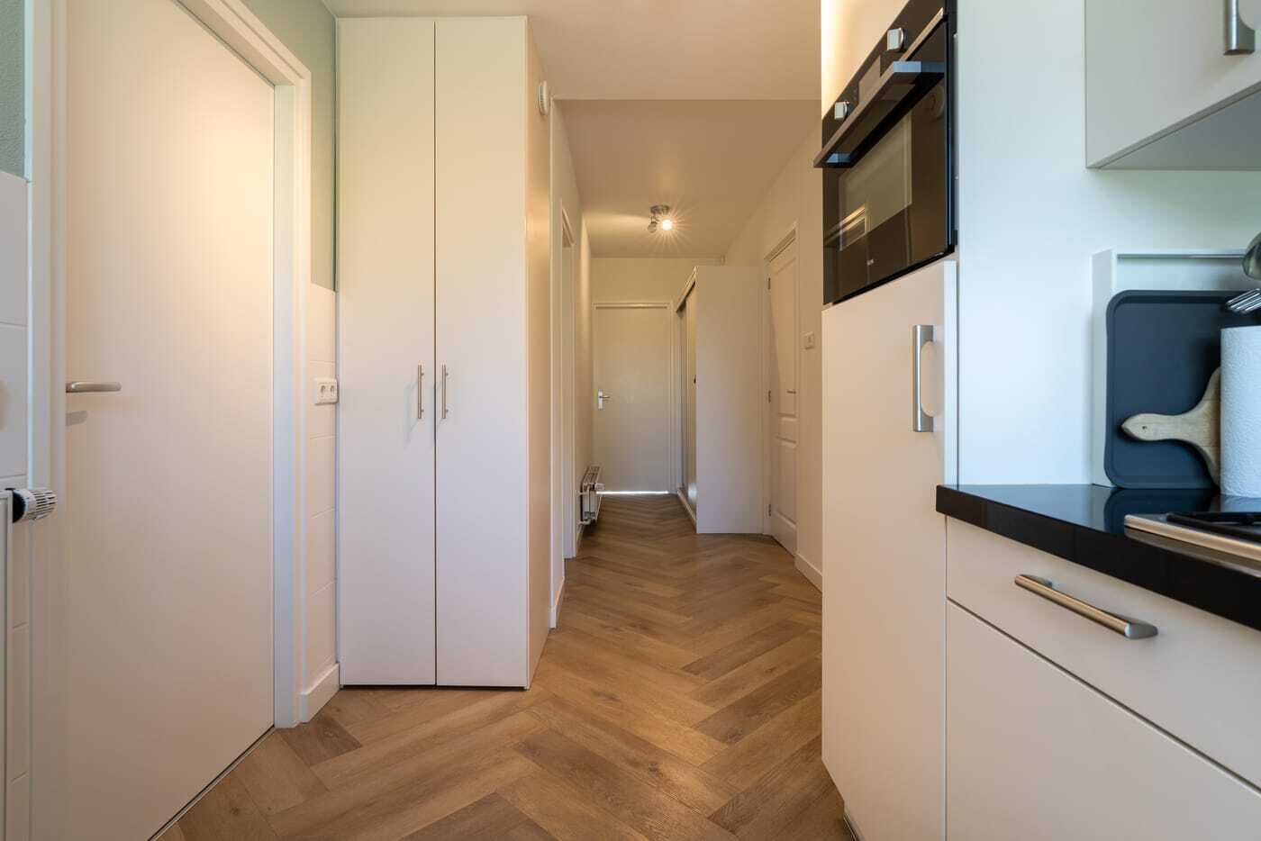 Ein schmaler Flur mit Holzboden, weißen Schränken und einer Küchenzeile mit einer Mikrowelle und einem Schneidebrett.
