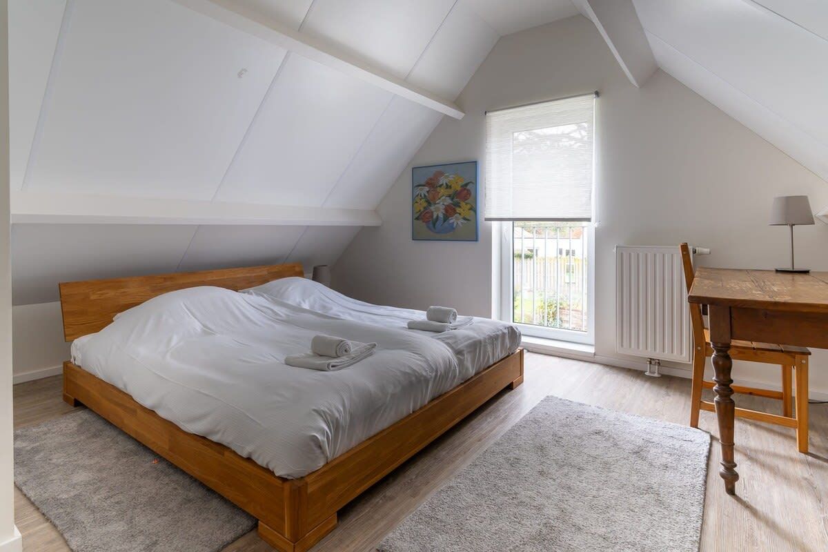 Un dormitorio con cama de madera, ventana con persianas, radiador, mesa con lámpara y un cuadro en la pared.