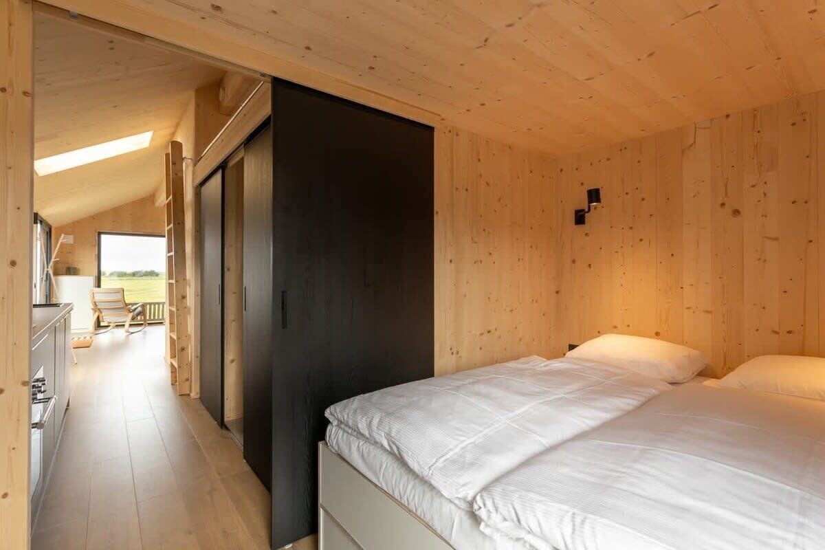 A wooden interior with a bed, a dark door, and a view of the outdoors.