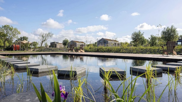 A serene outdoor pool area with a clear sky, surrounded by greenery and modern buildings. - Cosy tiny house for two in Westkapelle | Zeeuwse Liefde – Tiny House 3 (Westkapelle)