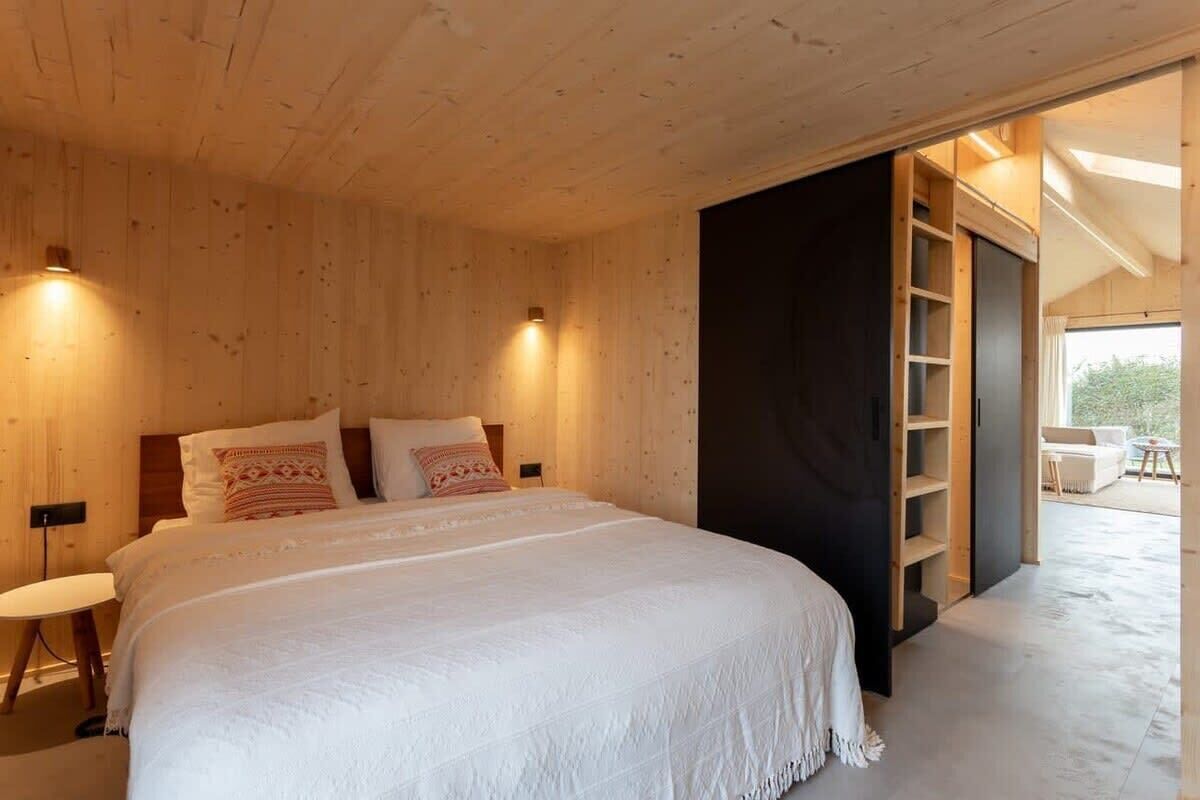 Een slaapkamer met een houten plafond, een bed met wit beddengoed en een donkere wand met een inbouwkast.