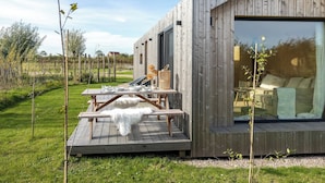 A modern wooden cabin with a deck, large windows, and a view of the surrounding landscape. - Cosy tiny house for two in Westkapelle | Zeeuwse Liefde – Tiny House 3 (Westkapelle)