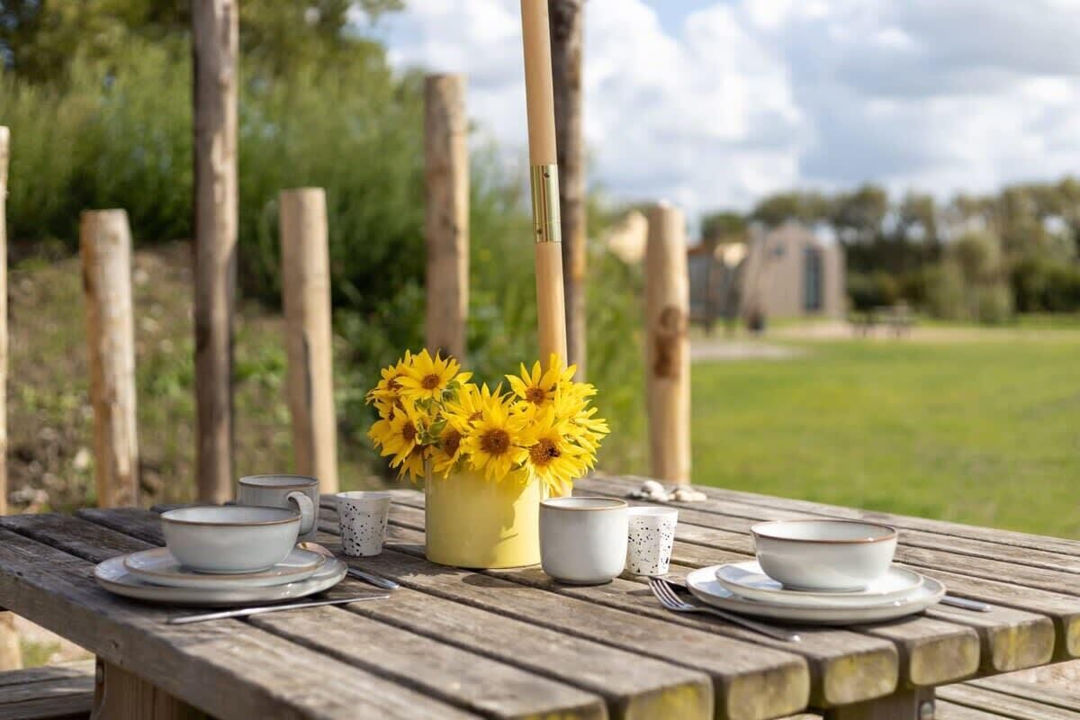 A wooden picnic table with a vase of yellow flowers, white cups, and saucers.