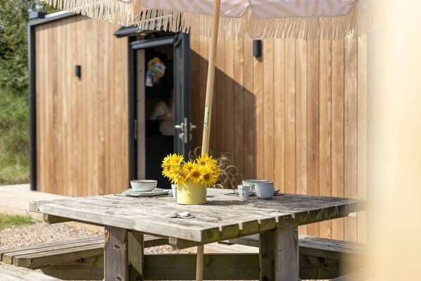 A wooden outdoor table with a vase of yellow flowers, a bench, and a wooden structure with a door in the background.
