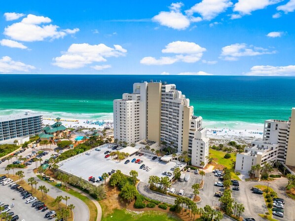 A coastal hotel with multiple high-rise buildings, a parking lot, and a beach in the background.