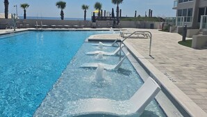 A swimming pool with a water feature, surrounded by a tiled deck and palm trees, with a building in the background.