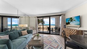 A modern hotel room with a large balcony, a sofa, a dining table, and a TV.