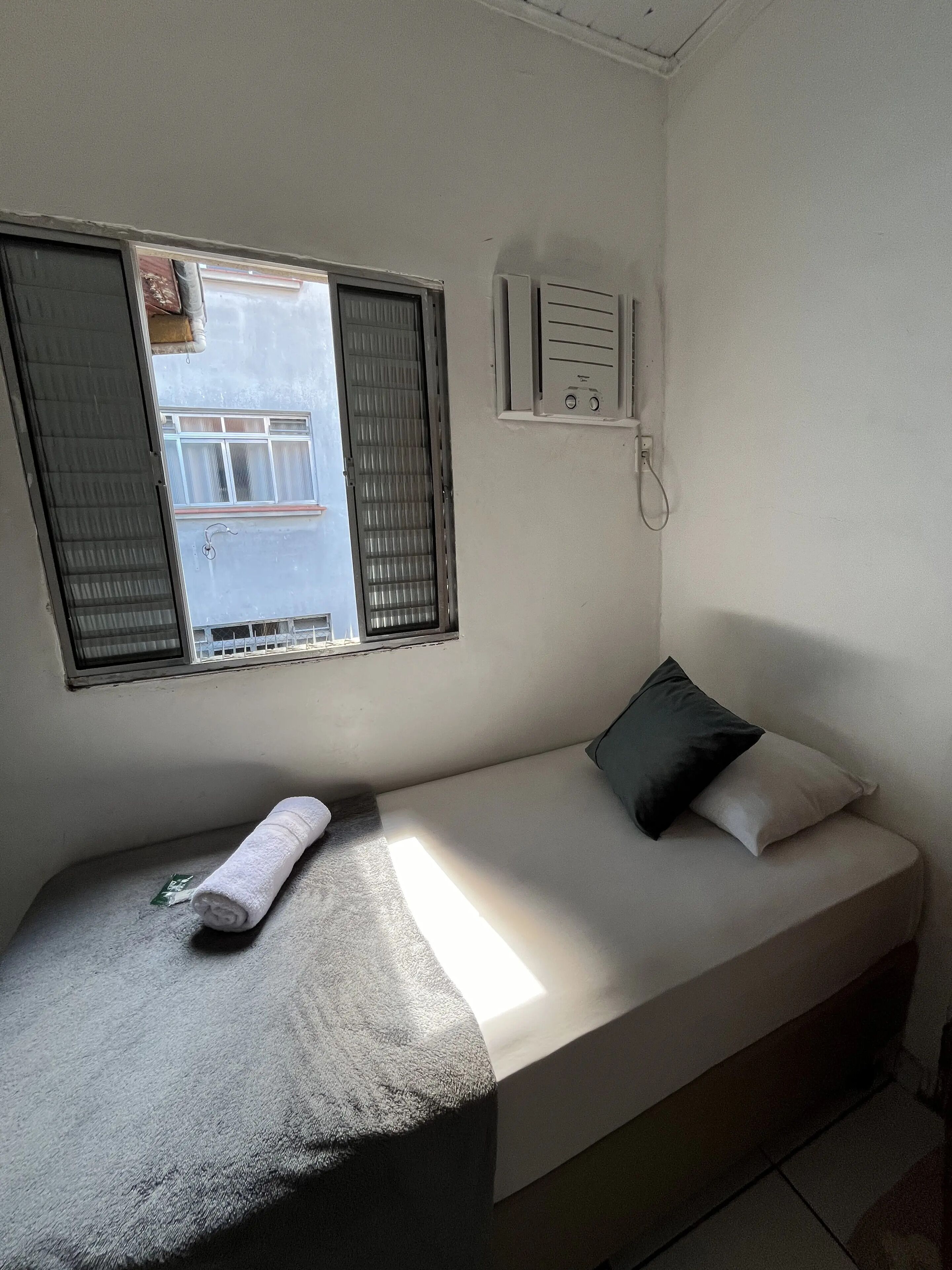A small, single-bed room with a window, a power outlet, and a towel on the bed.