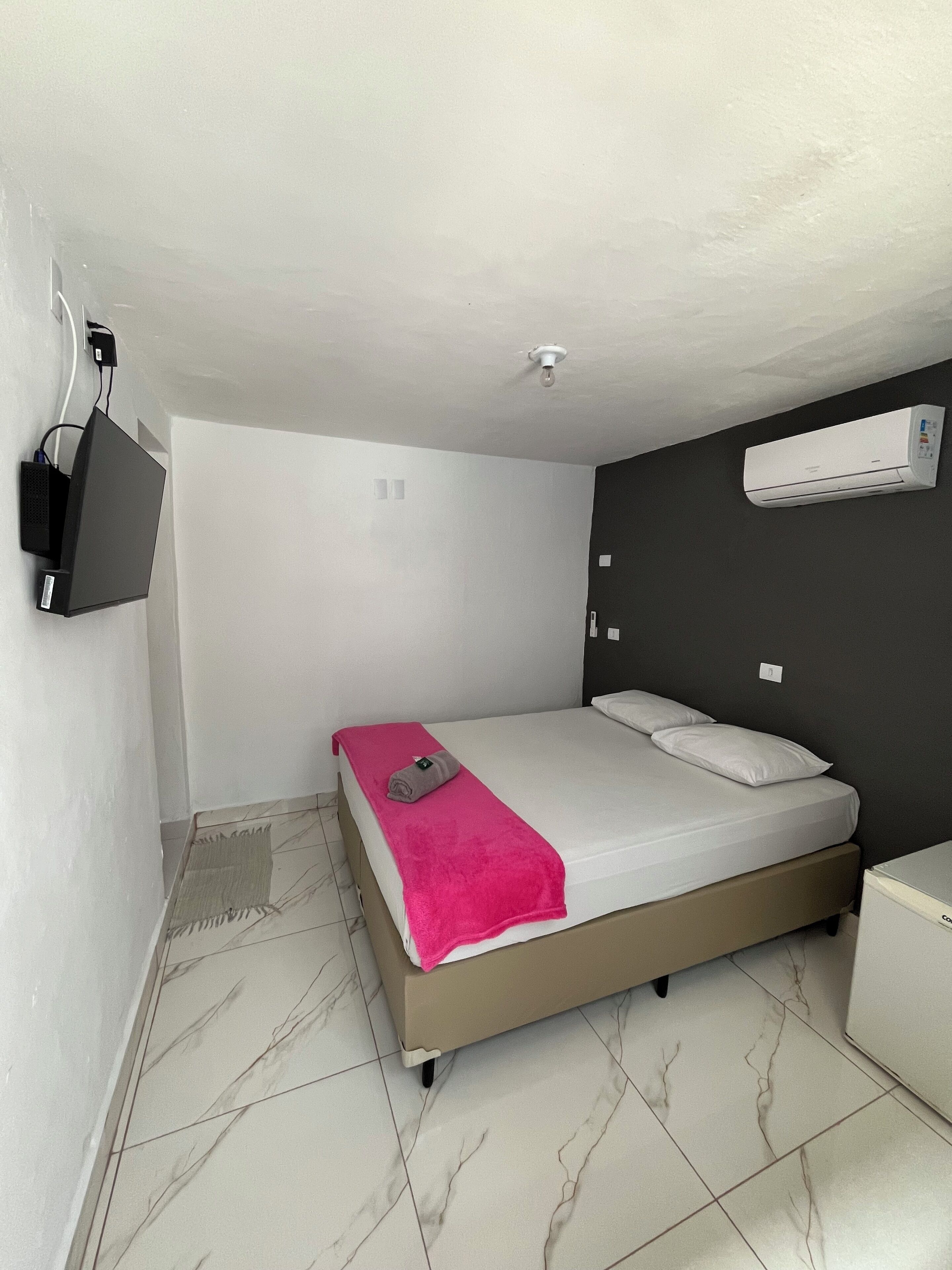 A small, modern bedroom with a bed, a TV mounted on the wall, and an air conditioning unit.