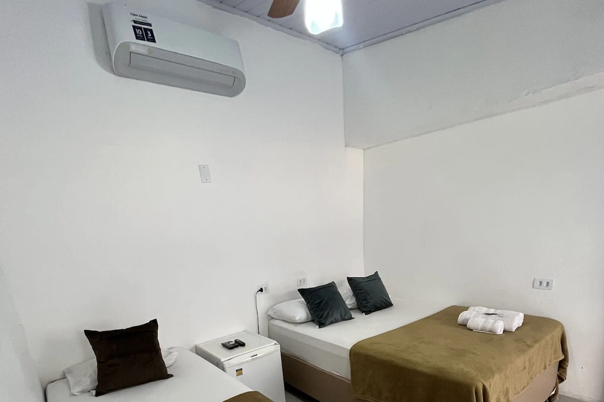 Habitación de hotel con dos camas, cada una con una manta marrón y una toalla enrollada. Se observa un ventilador de techo, un equipo de aire acondicionado y una mesita de noche pequeña con una lámpara.