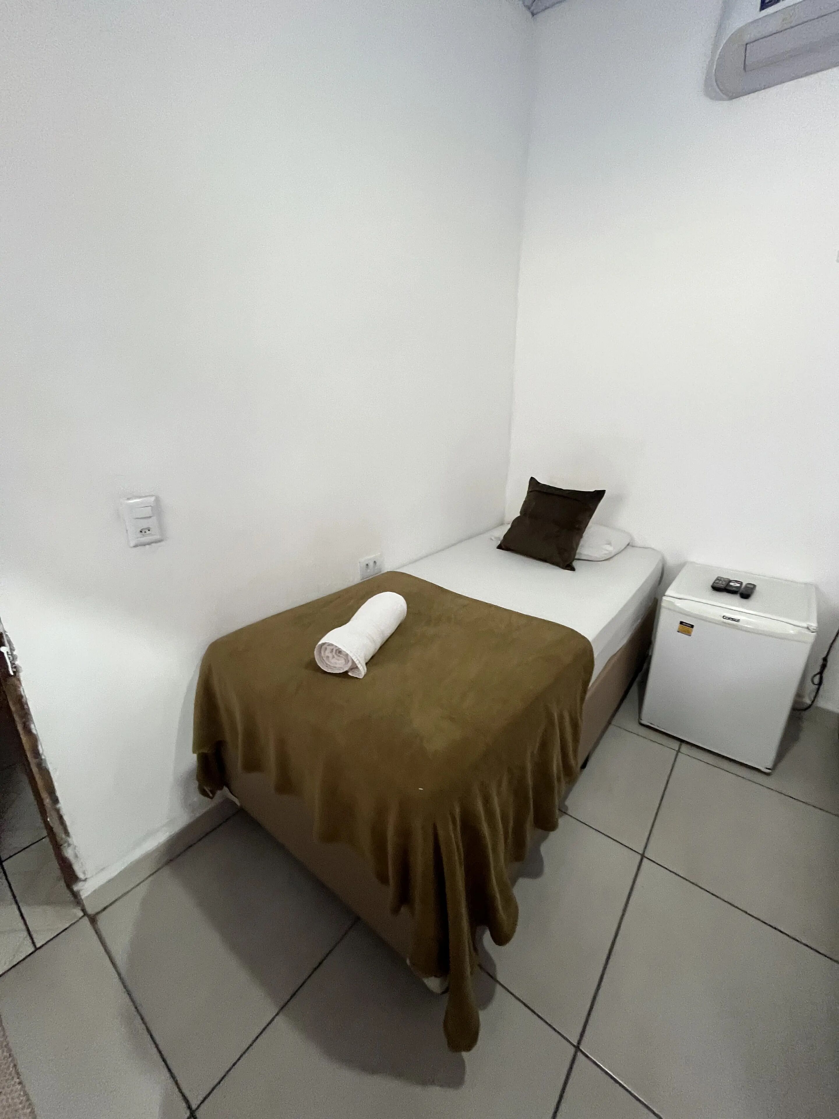 Una habitación pequeña con cama individual, cubrecama marrón, almohada blanca, mesita de noche blanca de tamaño reducido con teléfono y control remoto, y un calefactor blanco compacto.