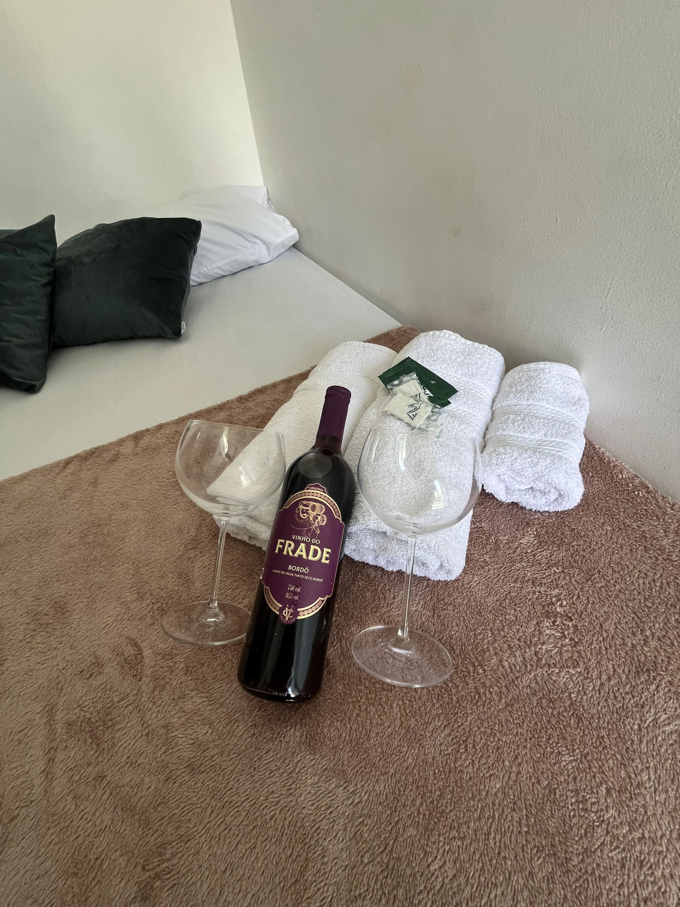 En vinflaska, ett vinglas och hopvikta handdukar på en brun matta i ett rum med en vit säng och en mörk kudde.