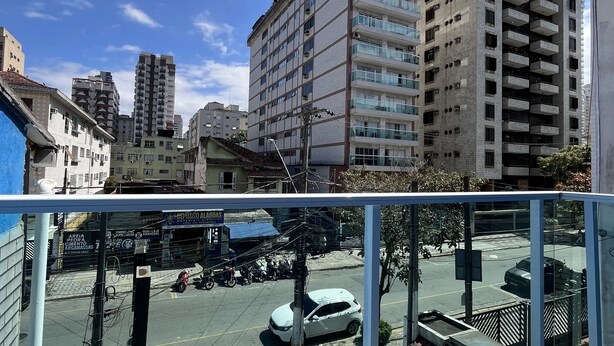 A high-rise building with multiple floors and balconies, a street with parked cars and a few pedestrians, and a clear blue sky. - Pousada Aloha Santos (Santos)