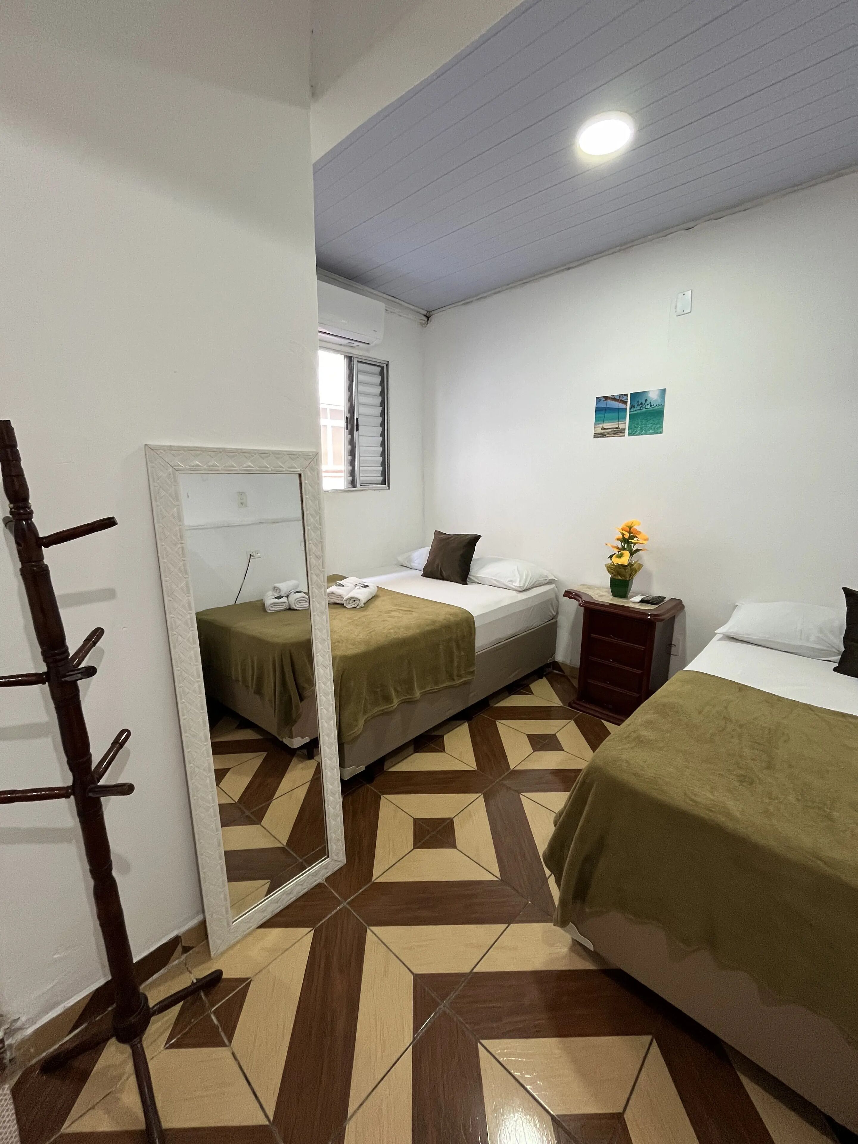A hotel room with two beds, a mirror, a wooden chair, a small table with a vase, and a tiled floor.