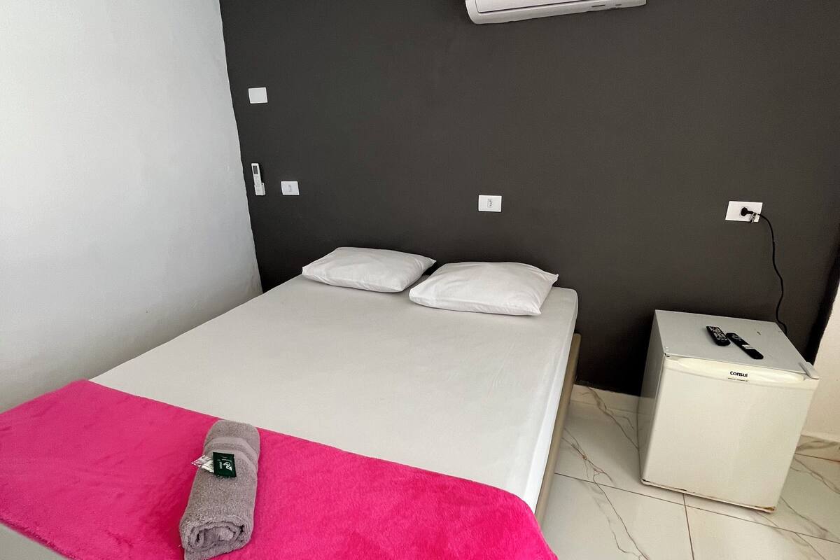 Un dormitorio moderno con una cama, una toalla rosa brillante, un aire acondicionado y un armario blanco pequeño.