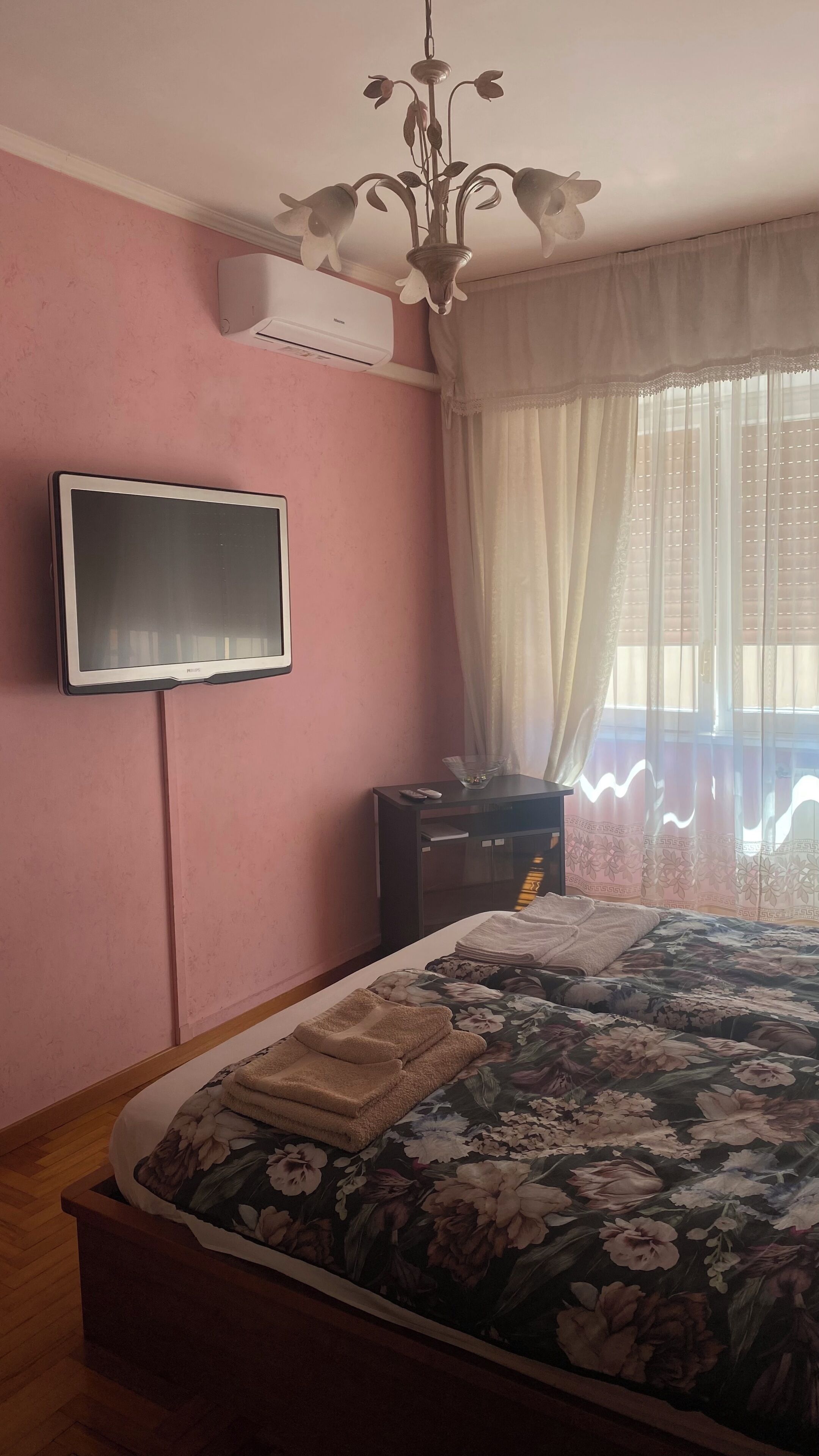 Un dormitorio con cama, televisión montada en la pared, mesita de noche con control remoto y ventana con cortinas translúcidas.