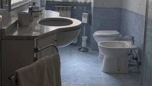 Une salle de bain moderne dotée d’un grand lavabo, de toilettes et d’un bidet. Elle comprend un grand miroir, une fenêtre avec des rideaux à motifs et des murs carrelés.