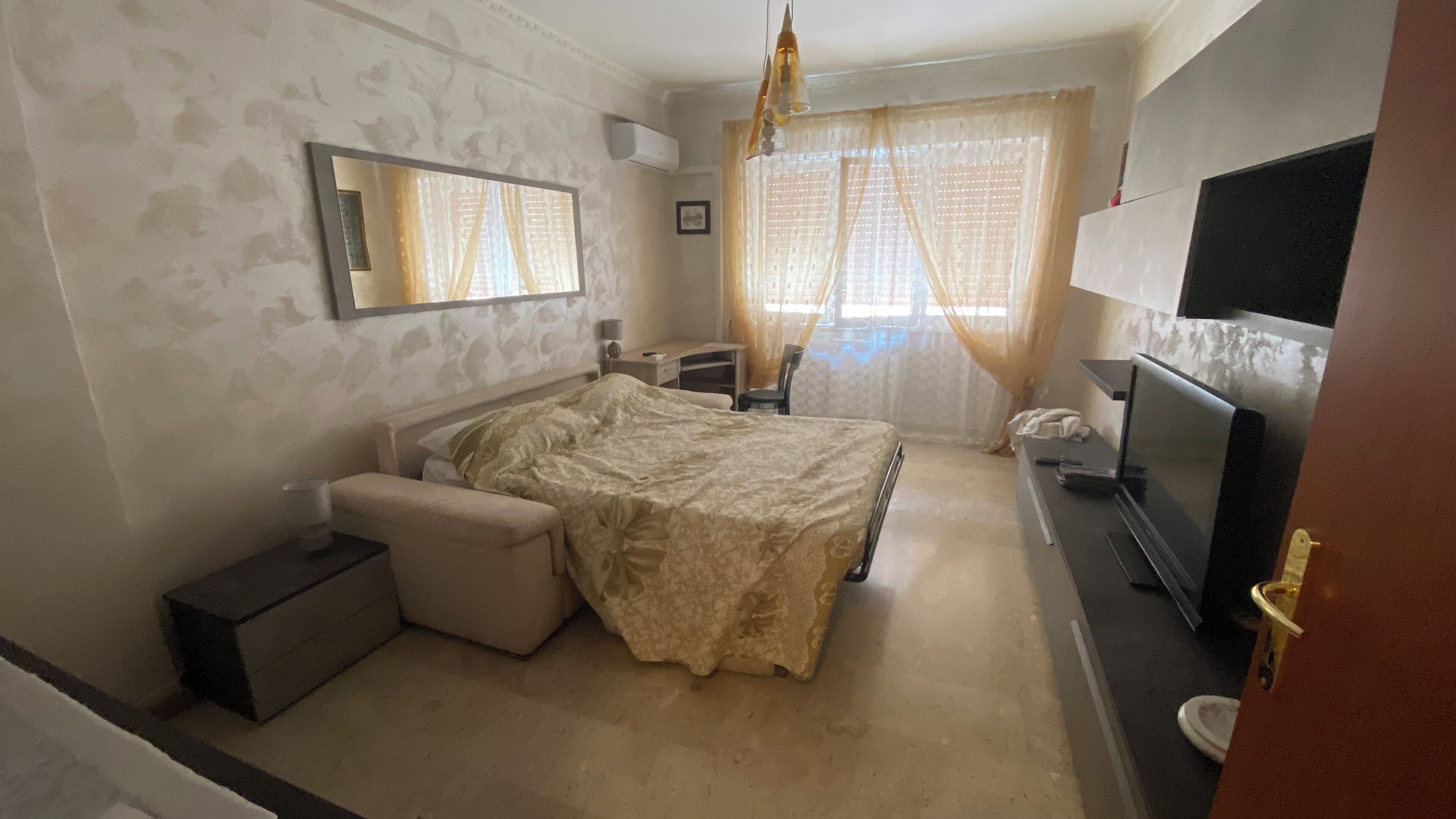 Ein Schlafzimmer mit Bett, Nachttisch, Stuhl, Fernseher und Vorhängen.