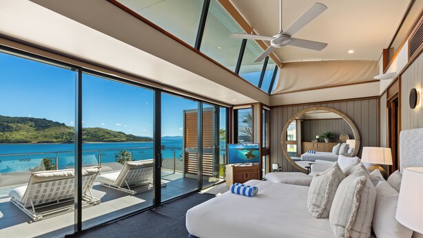 A spacious balcony with a clear view of the sea and hills, a large bed with multiple pillows, and a ceiling fan.