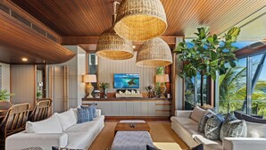 A modern living room with a sofa set, a coffee table, and a TV. There are wicker lamps, a plant, and a view of the outdoors.