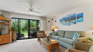 A spacious living room with a glass coffee table, a sofa, and a chair. There is a ceiling fan, a large window with curtains, and a door leading to an outdoor area.